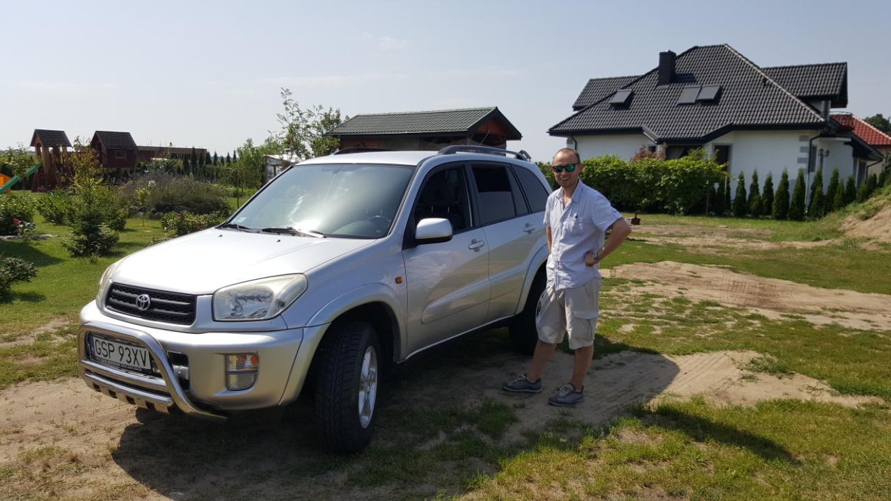 Srebrny SUV Toyota RAV4 z relingami dachowymi i chromowanym orurowaniem przednim, zaparkowany na trawniku obok mężczyzny w okularach przeciwsłonecznych, z domem jednorodzinnym w tle.