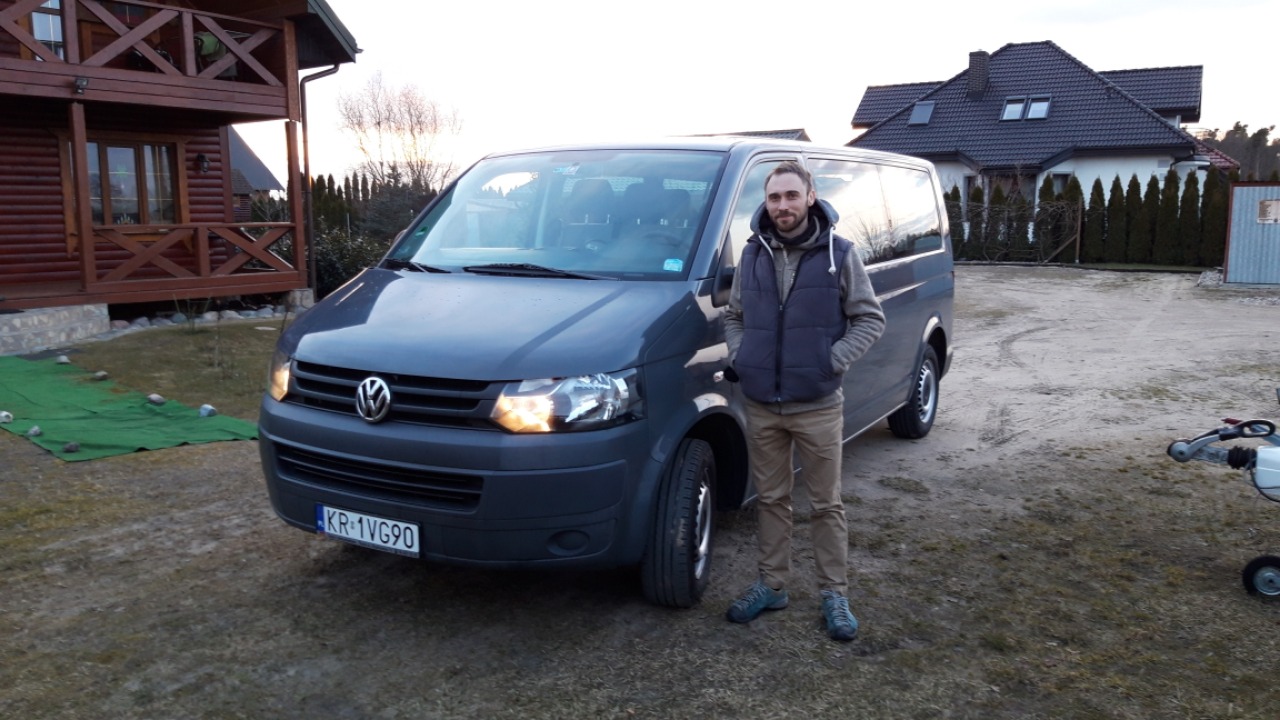 Szary bus Volkswagen Transporter z kierowcą na tle domu z bali i przyczepy, tablica rejestracyjna KR-1VG90.