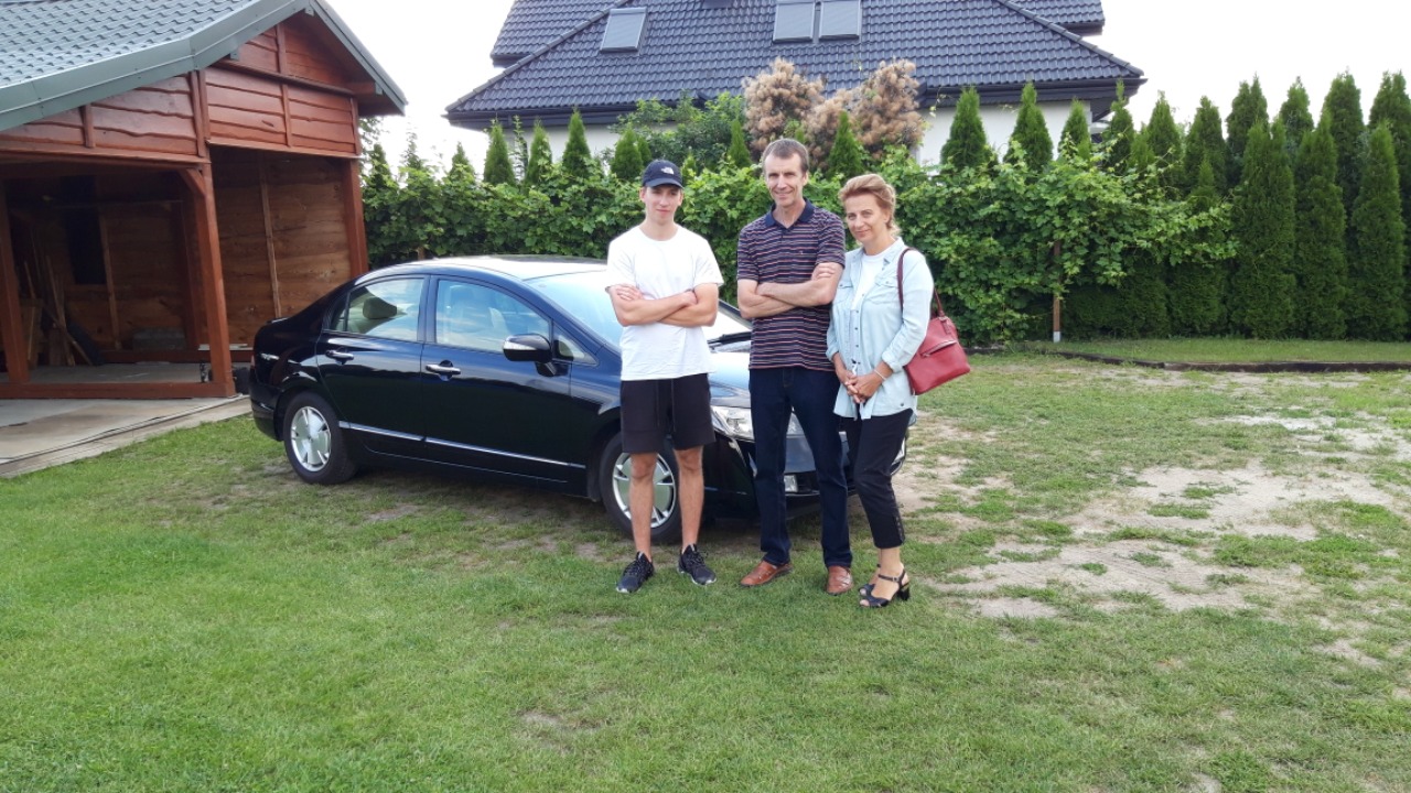 Rodzinne zdjęcie z czarnym sedanem na trawniku przed domem z drewnianą wiatą garażową.