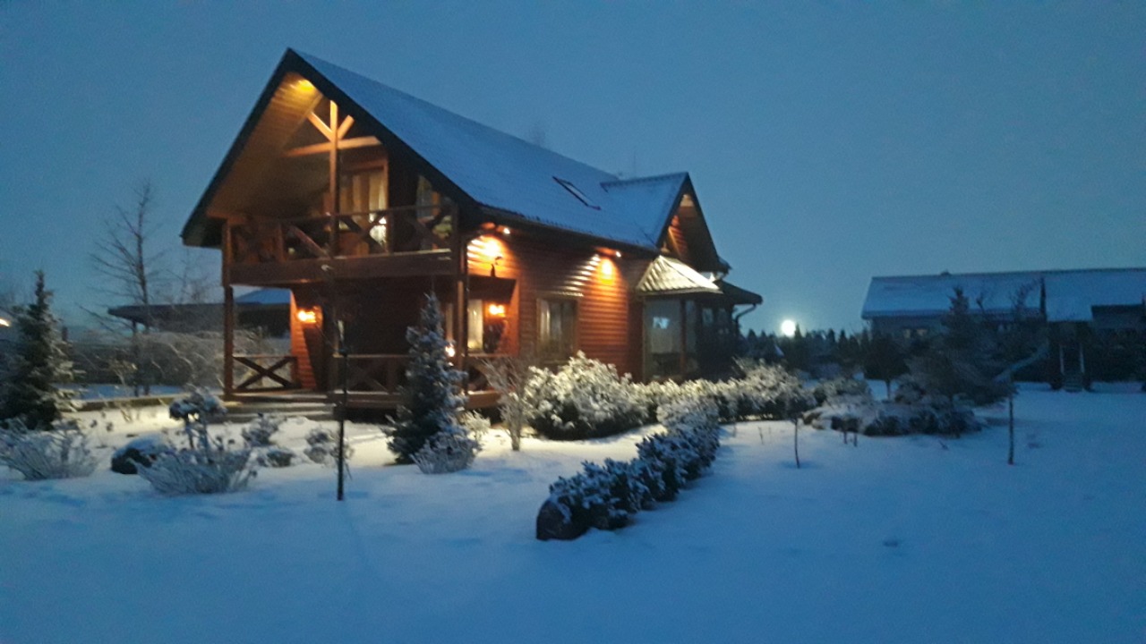 Oświetlony, drewniany dom z poddaszem użytkowym i balkonem, otoczony śniegiem i zimozielonymi krzewami w zimowej scenerii o zmierzchu.