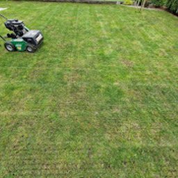 Wojciech Palkij Firma Handlowo-Usługowa - Aeracja trawnika: mężczyzna w czarnych spodniach i szarej bluzie obsługuje zielony aerator spalinowy na tle równomiernie nakłutego trawnika.