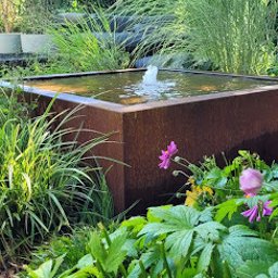Wojciech Palkij Firma Handlowo-Usługowa - Rustykalny, kwadratowy zbiornik wodny z fontanną w bujnym ogrodzie. Rośliny o dużych liściach i wysokie trawy ozdobne tworzą naturalne tło.