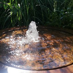 Wojciech Palkij Firma Handlowo-Usługowa - Oczko wodne z fontanną w rustykalnym stylu, otoczone zielenią. Woda tryska pionowo w górę, tworząc mały strumień. Widoczne refleksy światła na powierzchni wody.