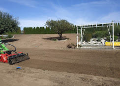 Czerwony traktor przygotowuje glebę pod trawnik na tle zieleni i białej konstrukcji. Widok na równą, brązową powierzchnię ziemi po obróbce.