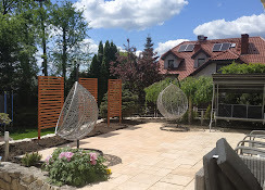 Ukończony taras z jasnych płyt, z dwoma wiszącymi fotelami w kształcie łezki i drewnianymi panelami. W tle zadbany ogród i dom z czerwonym dachem.