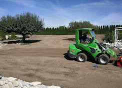 Zielona miniładowarka Avant przygotowuje teren pod trawnik, widoczny wyrównany grunt, drzewo i żywopłot w tle, błękitne niebo.