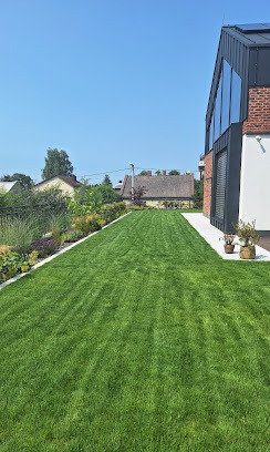 Perfekcyjnie skoszony trawnik przed nowoczesnym domem z ceglaną elewacją i dużymi oknami. Widoczne rabaty kwiatowe i krzewy wzdłuż trawnika. Idealny trawnik.