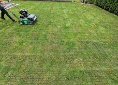 Aeracja trawnika: mężczyzna w czarnych spodniach i szarej bluzie obsługuje zielony aerator spalinowy na tle równomiernie nakłutego trawnika.