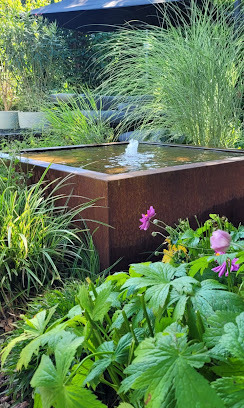 Rustykalny, kwadratowy zbiornik wodny z fontanną w bujnym ogrodzie. Rośliny o dużych liściach i wysokie trawy ozdobne tworzą naturalne tło.