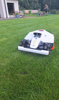 Biały robot koszący trawę na idealnie przystrzyżonym trawniku. W tle osoba z kosiarką spalinową i nowoczesny dom z białymi ścianami.