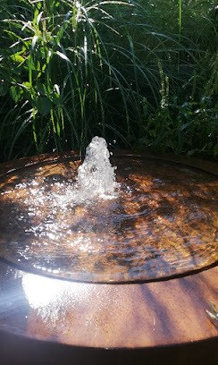 Oczko wodne z fontanną w rustykalnym stylu, otoczone zielenią. Woda tryska pionowo w górę, tworząc mały strumień. Widoczne refleksy światła na powierzchni wody.