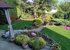 Zadbany ogród w Dobrej: równo skoszony trawnik, ozdobne krzewy i kwiaty, widok z perspektywy tarasu. Elementy małej architektury ogrodowej.