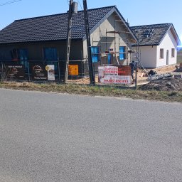 Firma Remontowo-Budowlana Grzegorz Konopka - Dwa domy w trakcie budowy, jeden z ocieploną elewacją i rusztowaniem, drugi w stanie surowym, ogrodzone siatką z reklamami firm budowlanych.
