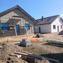 Budynek w trakcie prac elewacyjnych: szare ściany z rusztowaniem, okna zabezpieczone folią, drugi budynek z białą elewacją w tle, materiały budowlane na placu budowy.