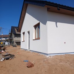 Firma Remontowo-Budowlana Grzegorz Konopka - Nowo otynkowana, biała elewacja domu jednorodzinnego z brązowymi oknami, widok z zewnątrz na plac budowy z piaskiem i materiałami budowlanymi.