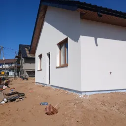 Nowo otynkowana, biała elewacja domu jednorodzinnego z brązowymi oknami, widok z zewnątrz na plac budowy z piaskiem i materiałami budowlanymi.