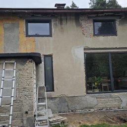 Firma Remontowo-Budowlana Grzegorz Konopka - Dwukondygnacyjny budynek w trakcie prac elewacyjnych, z widocznymi fragmentami starej elewacji, nowymi oknami w grafitowych ramach, dwiema aluminiowymi drabinami opartymi o ścianę i przygotowanym...