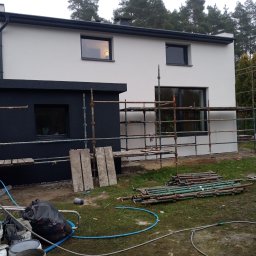 Firma Remontowo-Budowlana Grzegorz Konopka - Dwukondygnacyjny dom z elewacją w trakcie wykonywania, widoczne rusztowania, część elewacji pomalowana na biało, część na czarno, na trawniku leżą narzędzia i materiały budowlane.