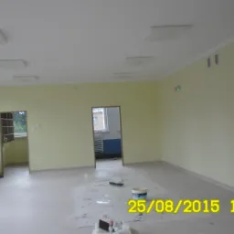 Jasne wnętrze po malowaniu ścian na kremowo, z widocznymi oknami, grzejnikami, częściowo zabezpieczoną podłogą i narzędziami malarskimi.