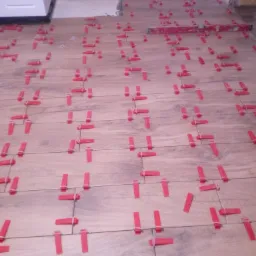 Ułożone płytki podłogowe imitujące drewno, z czerwonymi klipsami poziomującymi i poziomicą w tle, fragment pomieszczenia w trakcie prac wykończeniowych.