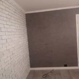 Wnętrze po remoncie: ściana z białej cegły dekoracyjnej, szara ściana z fakturą, szara podłoga imitująca drewno, biała listwa przypodłogowa i sufitowa, widoczny kabel na podłodze.