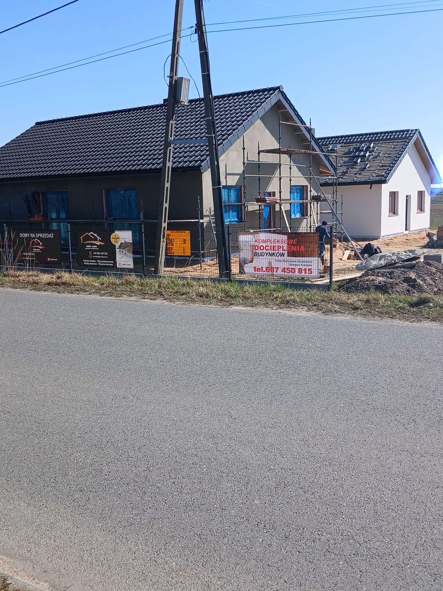 Dwa domy w trakcie budowy, jeden z ocieploną elewacją i rusztowaniem, drugi w stanie surowym, ogrodzone siatką z reklamami firm budowlanych.