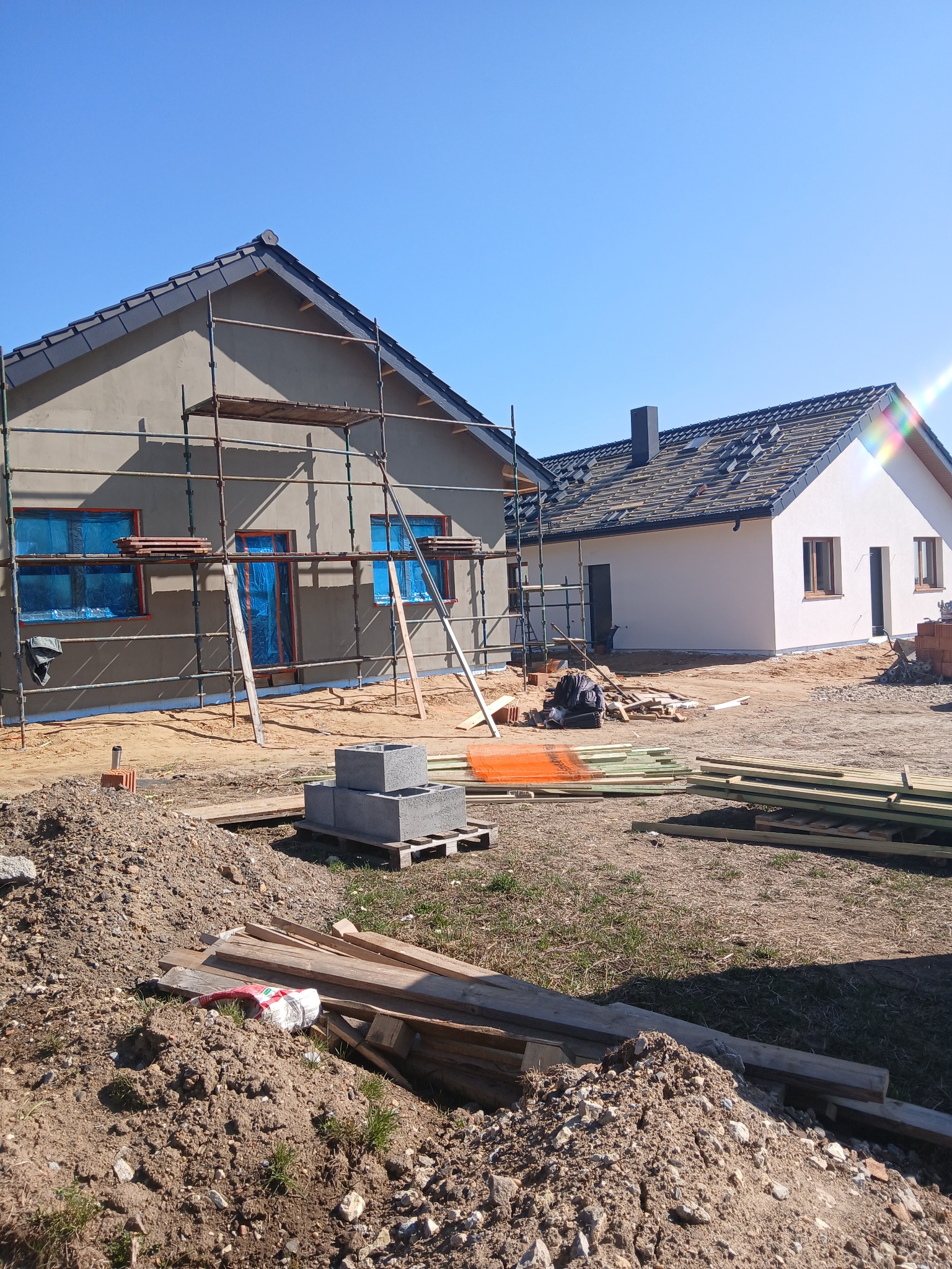 Budynek w trakcie prac elewacyjnych: szare ściany z rusztowaniem, okna zabezpieczone folią, drugi budynek z białą elewacją w tle, materiały budowlane na placu budowy.