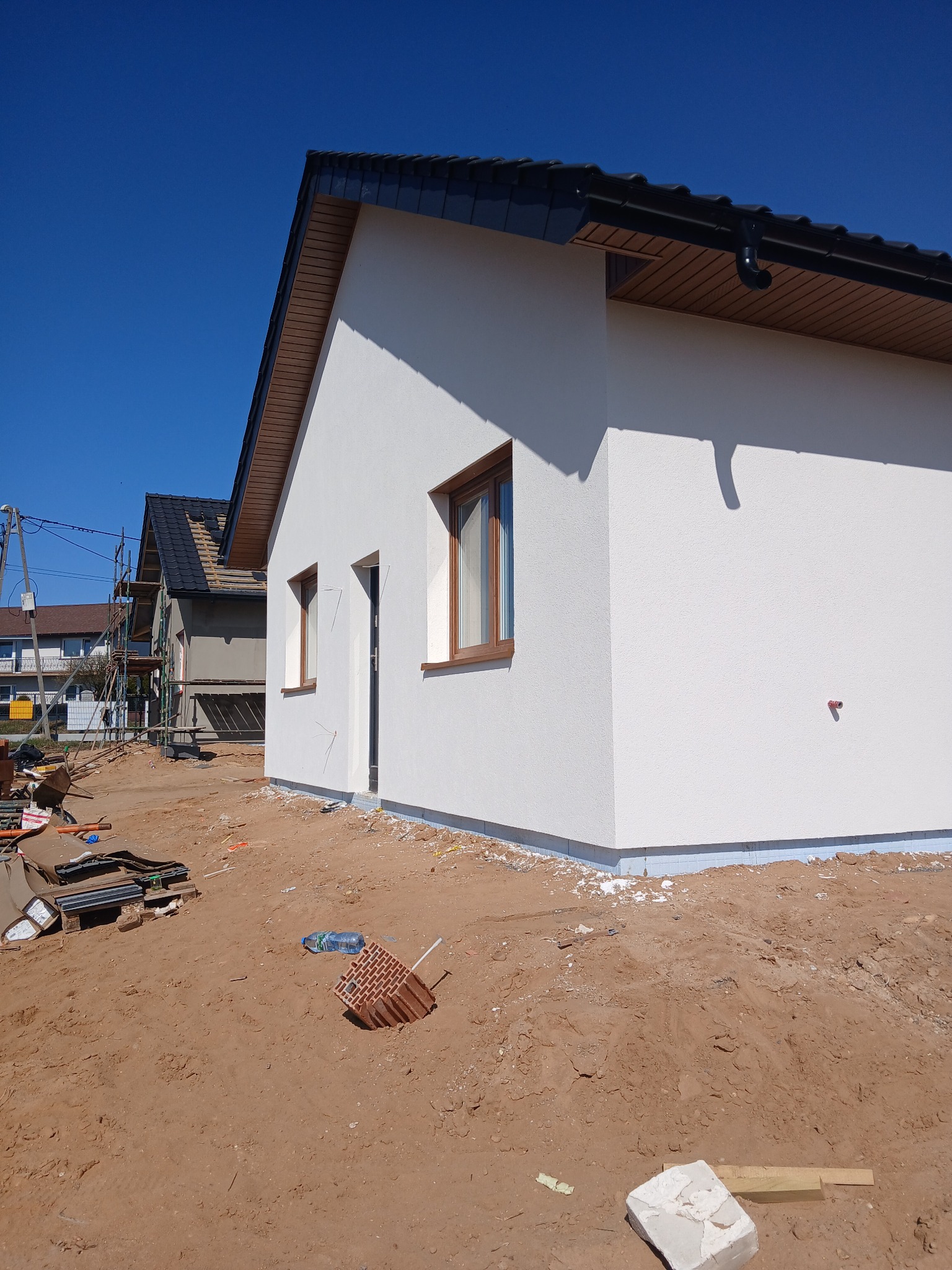 Nowo otynkowana, biała elewacja domu jednorodzinnego z brązowymi oknami, widok z zewnątrz na plac budowy z piaskiem i materiałami budowlanymi.