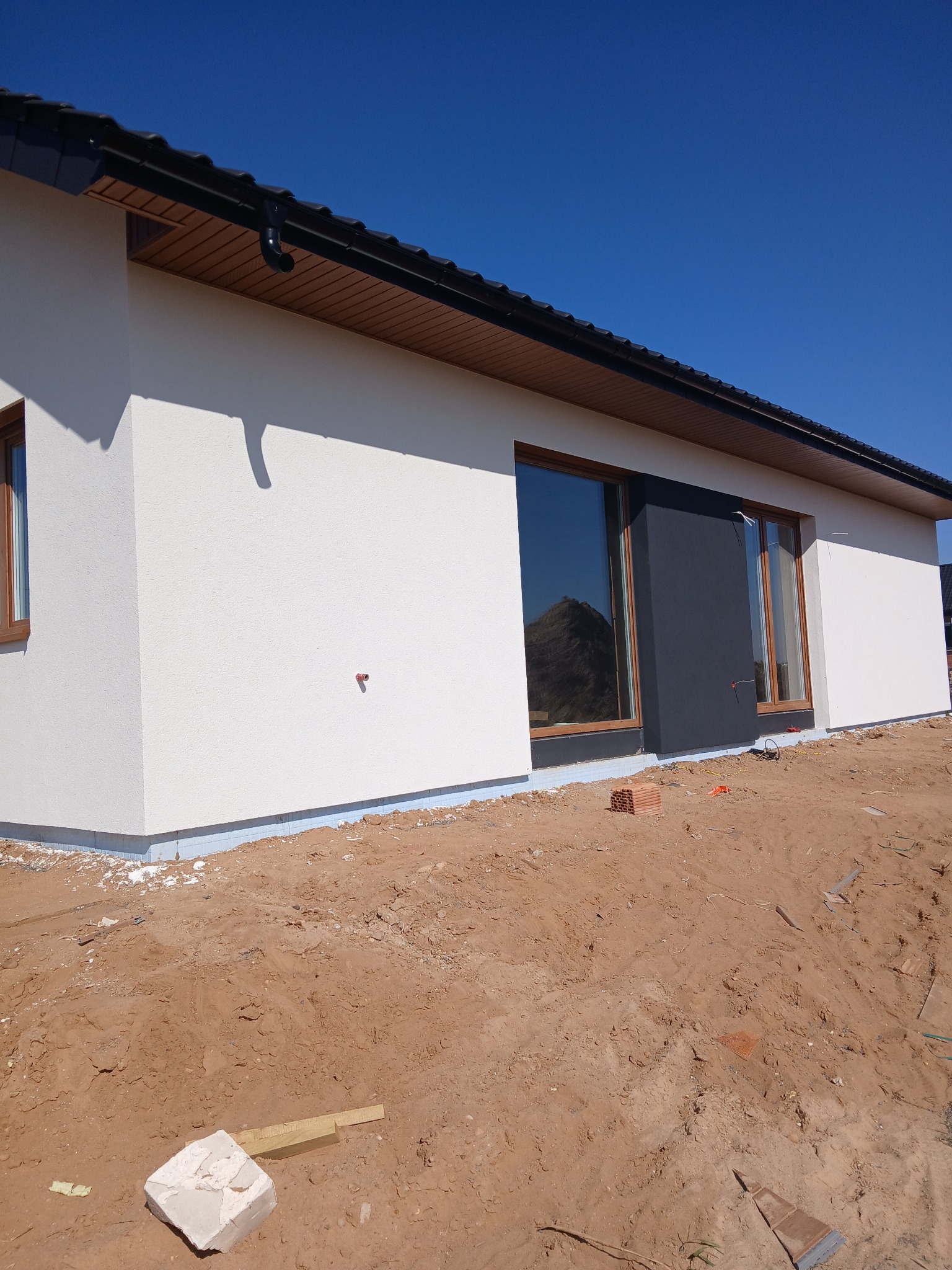 Fragment elewacji domu jednorodzinnego z oknami, częściowo otynkowana na biało, z fragmentem w kolorze grafitowym, widoczne przygotowania do dalszych prac wykończeniowych.