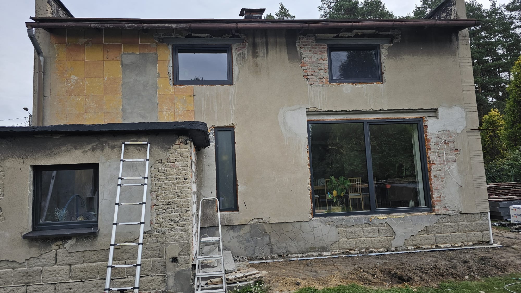 Dwukondygnacyjny budynek w trakcie prac elewacyjnych, z widocznymi fragmentami starej elewacji, nowymi oknami w grafitowych ramach, dwiema aluminiowymi drabinami opartymi o ścianę i przygotowanym...