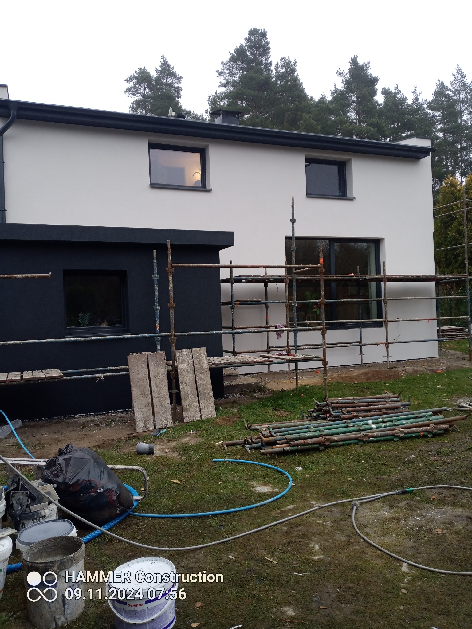 Dwukondygnacyjny dom z elewacją w trakcie wykonywania, widoczne rusztowania, część elewacji pomalowana na biało, część na czarno, na trawniku leżą narzędzia i materiały budowlane.