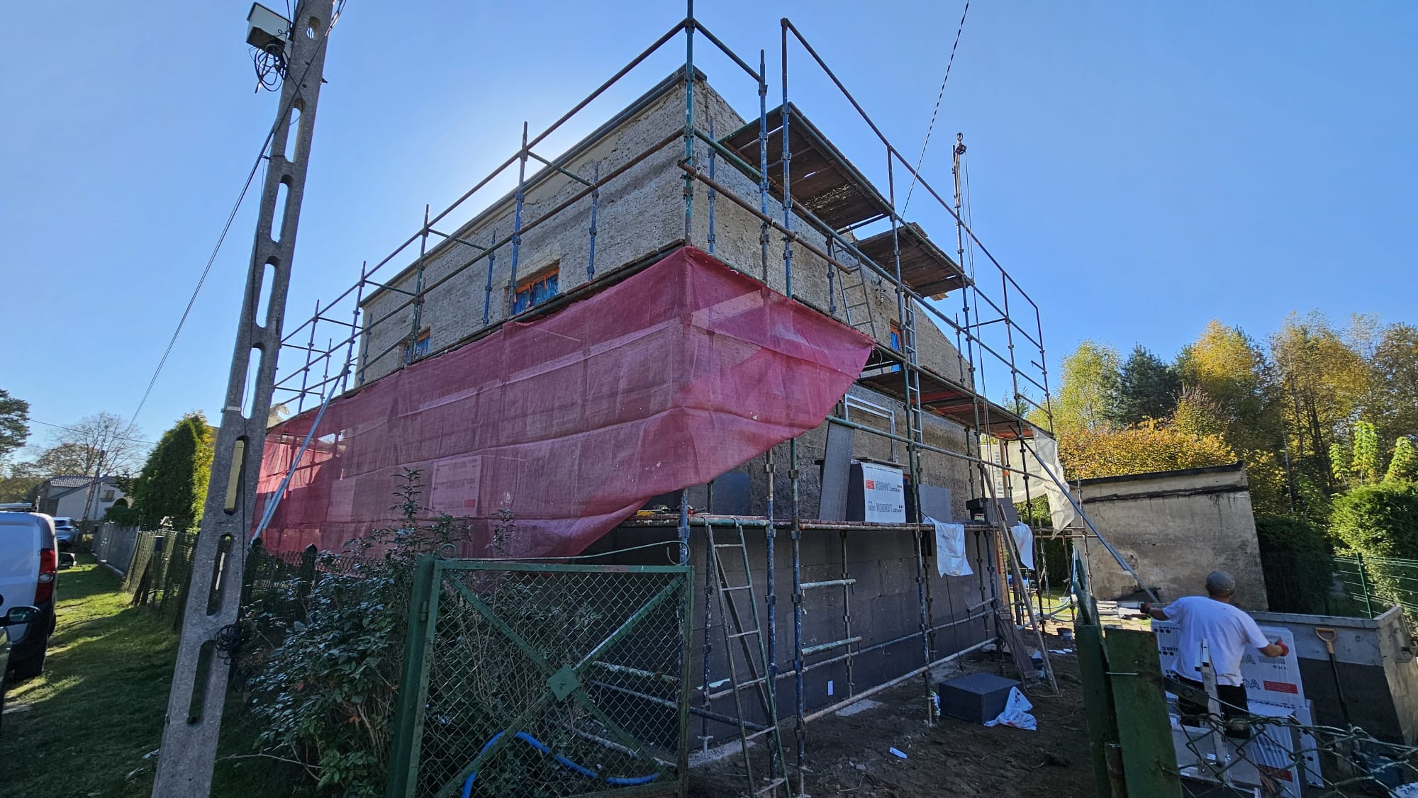 Dwupiętrowy budynek w trakcie prac elewacyjnych, otoczony rusztowaniami i częściowo osłonięty czerwoną siatką. Widoczne płyty izolacyjne na ścianie i pracownik w białej koszulce przy pracy.