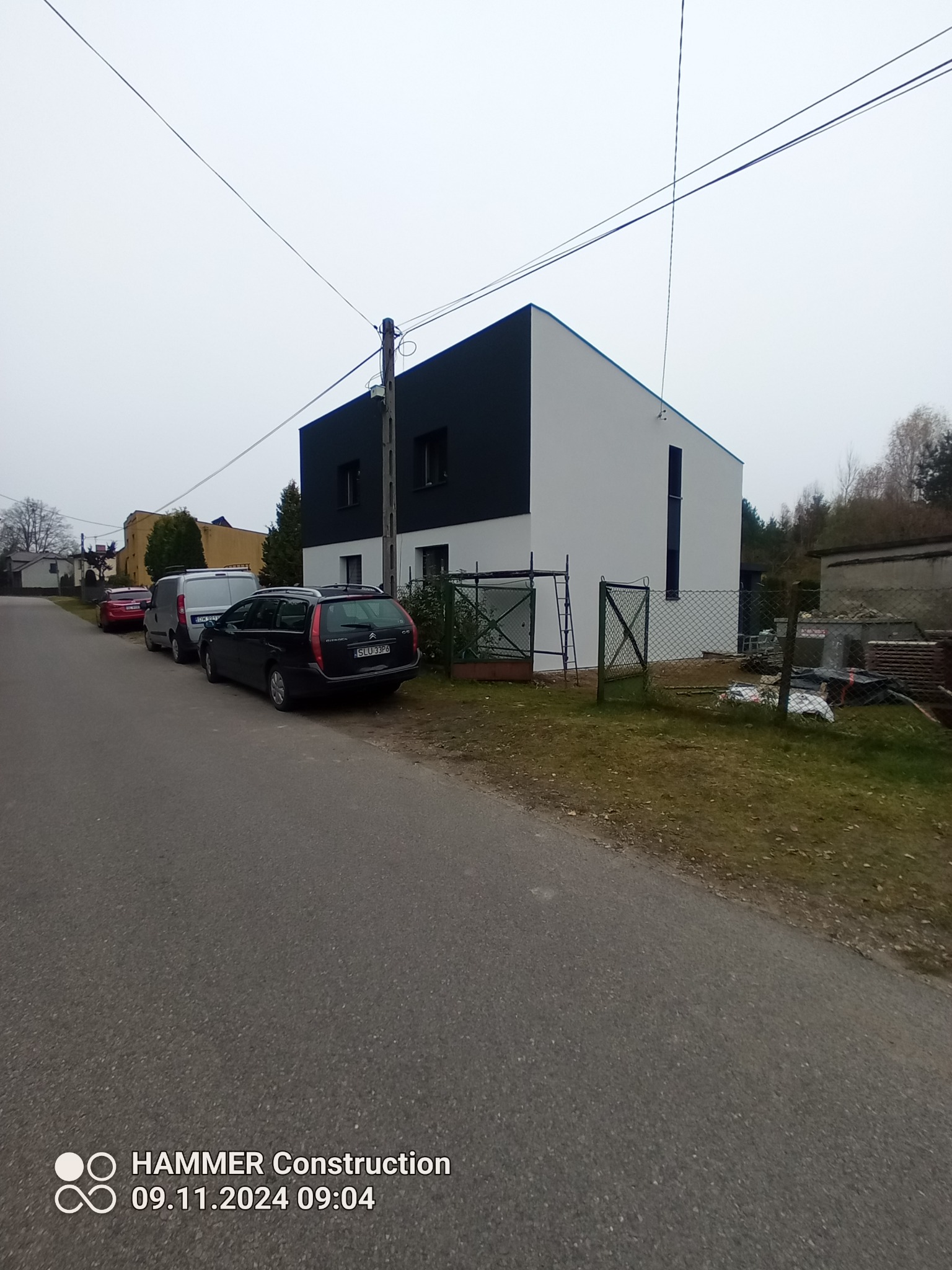 Nowoczesny dom jednorodzinny z czarno-białą elewacją, widoczny z ulicy, z zaparkowanymi samochodami i elementami ogrodzenia, w pochmurny dzień.