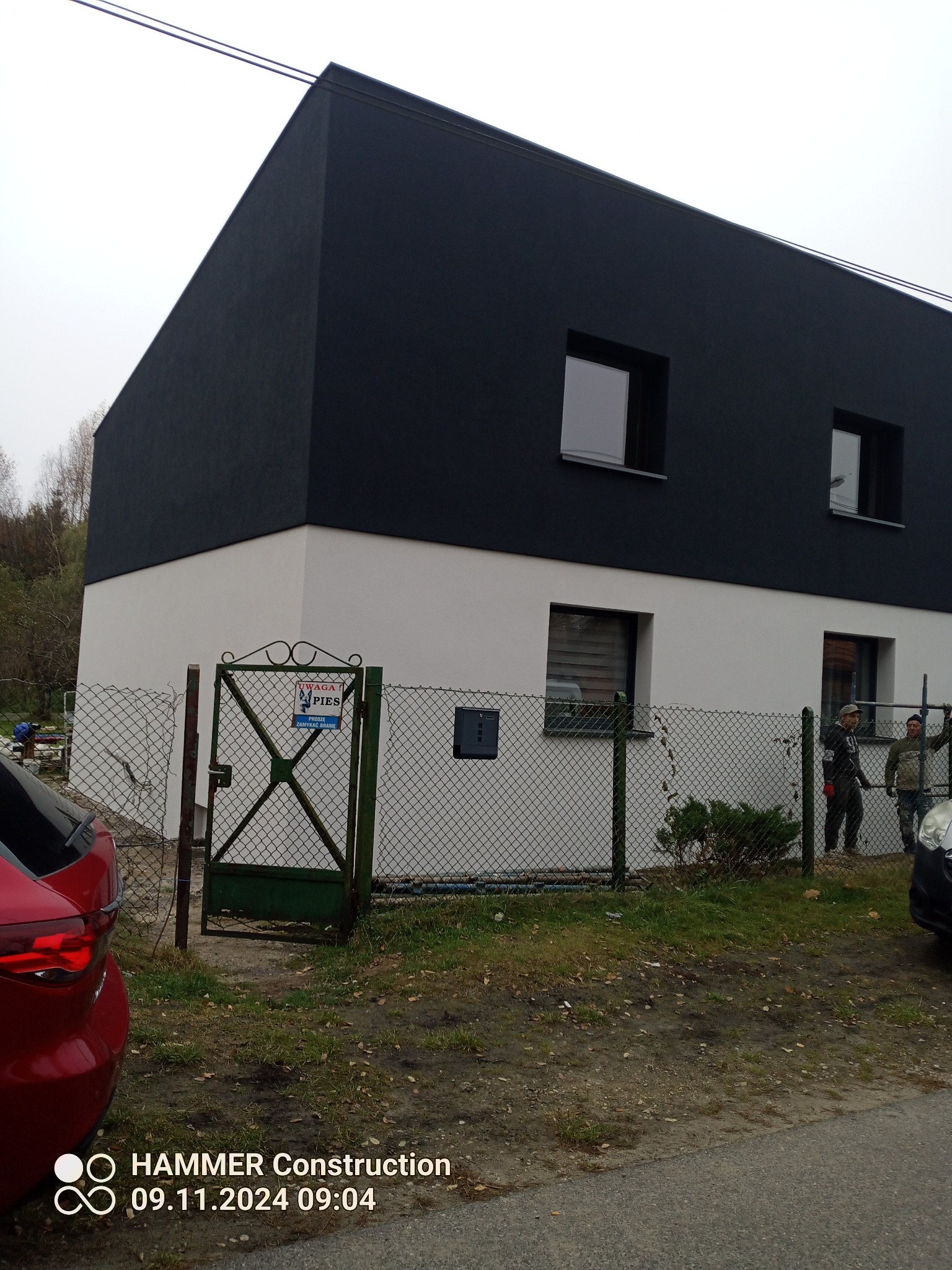 Nowoczesny dom z dwukolorową elewacją (biały dół, grafitowa góra) widziany zza ogrodzenia; dwóch pracowników przy rusztowaniu po prawej stronie.