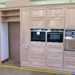 Niedokończona zabudowa kuchenna z jasnego drewna z wbudowanymi sprzętami AGD: ekspresem do kawy, piekarnikiem i otwartą zmywarką. Widoczna surowa aranżacja wnętrza warsztatu stolarskiego.