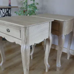 Dwa drewniane stoliki nocne w stylu ludwikowskim z szufladami i giętymi nogami, jeden z metalowym uchwytem, na drewnianej podłodze, w tle roślina doniczkowa i podstawa starej maszyny do szycia.