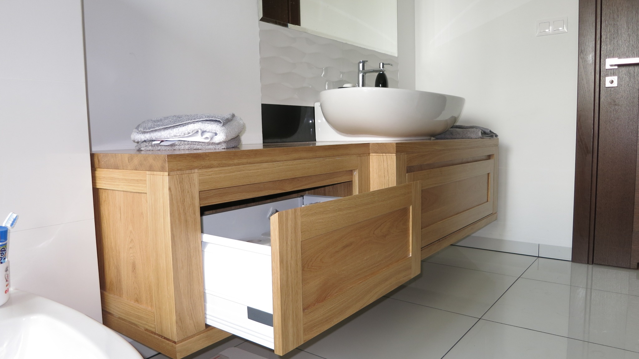 Nowoczesna łazienka z drewnianą szafką pod umywalkę z wysuwaną szufladą, białą umywalką nablatową i szarymi ręcznikami.