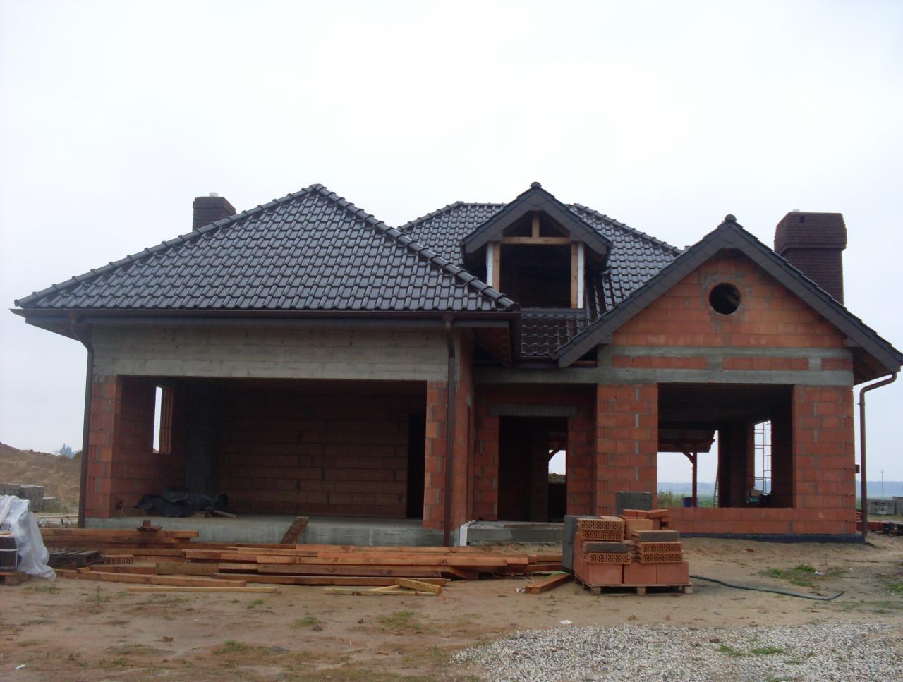 Dom w trakcie budowy z ciemną dachówką, widoczne cegły, drewno i rynny. Budynek dwukondygnacyjny z garażem, okna bez szyb, stan surowy otwarty.