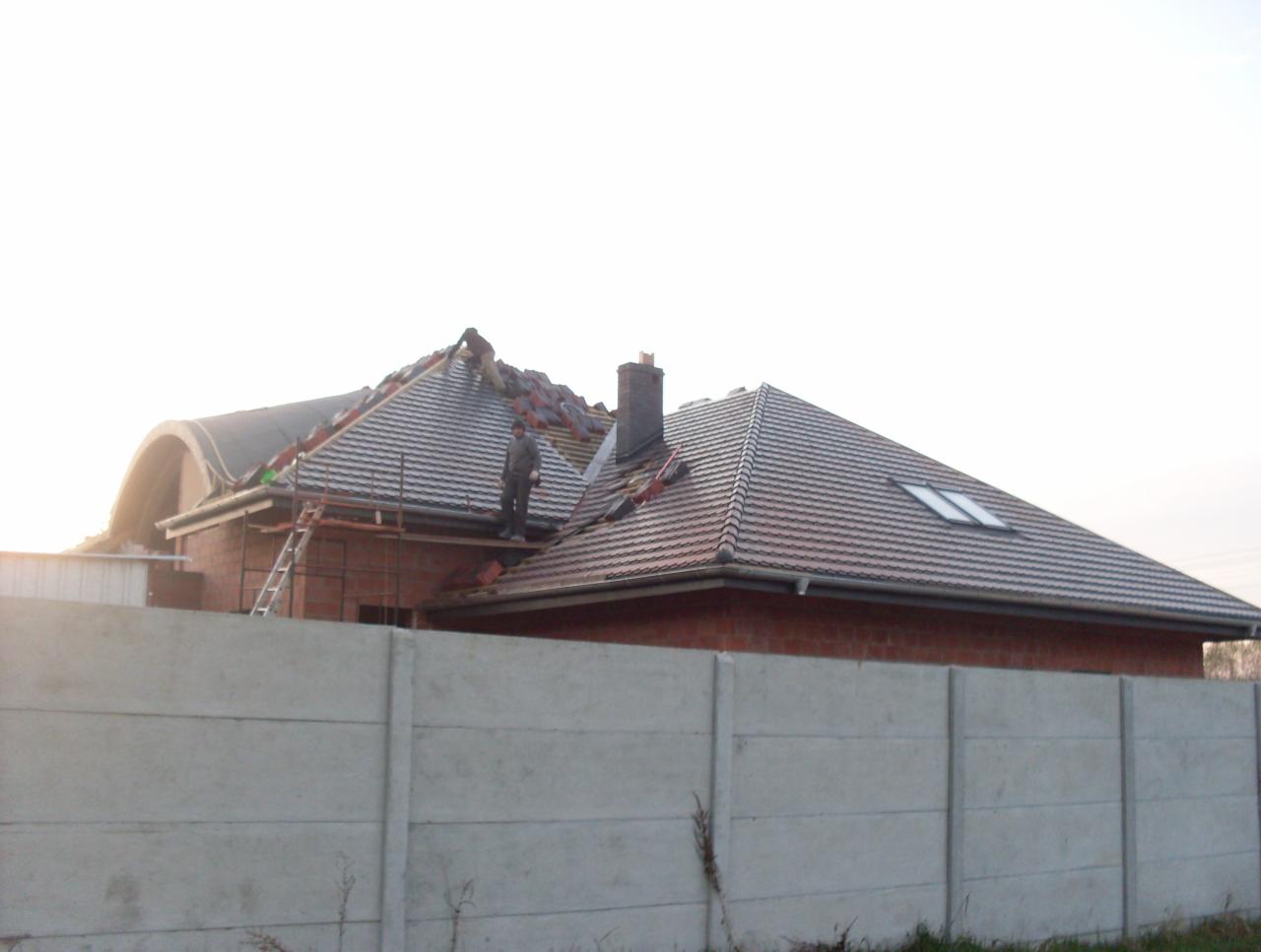 Ceglany dom w trakcie krycia dachu dachówką ceramiczną, widoczny pracownik na rusztowaniu i częściowo ułożona dachówka. W tle komin i okno dachowe.