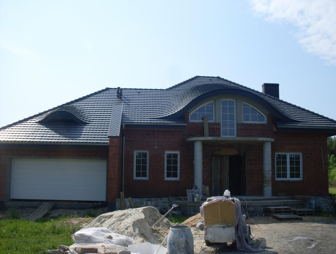 Dom jednorodzinny w trakcie budowy, z nowym, ciemnym dachem z dachówki ceramicznej. Widoczne materiały budowlane i mieszarka. Dach z oryginalnym oknem dachowym.