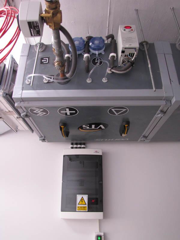 Szara centrala wentylacyjna VTS z podłączonymi przewodami i skrzynką elektryczną na białej ścianie. Widoczne elementy instalacji i oznaczenia bezpieczeństwa.