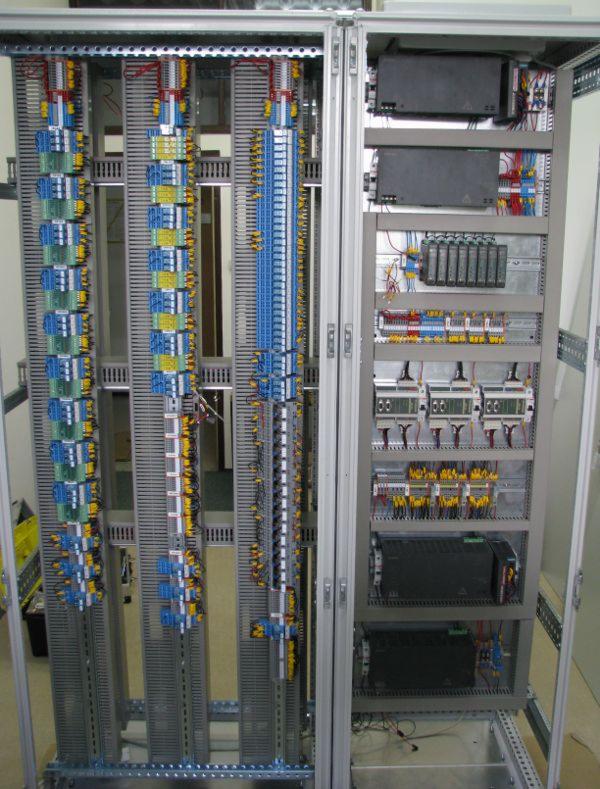 Otwarta szafa sterownicza z rozbudowaną instalacją elektryczną, widoczne liczne kable, złącza i moduły. Uporządkowany układ w szarej obudowie z elementami metalowymi.