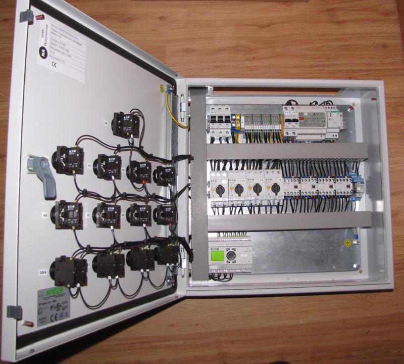 Otwarta szafa sterownicza z licznymi przekaźnikami, kablami i modułami, zamontowana na drewnianej powierzchni. Widoczne elementy automatyki i połączenia elektryczne.