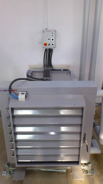 Szara centrala wentylacyjna Juwent z podłączonymi przewodami elektrycznymi i panelem sterowania na ścianie. Widoczne rury wentylacyjne w tle.