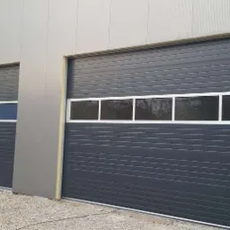 Dwie szare bramy garażowe segmentowe z oknami, zamontowane w szarej hali przemysłowej. Widok z zewnątrz na żwirowe podłoże.