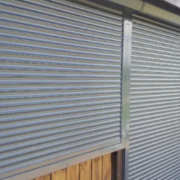 Szare, poziome, metalowe zabezpieczenie okna z widoczną butelką wody w tle.