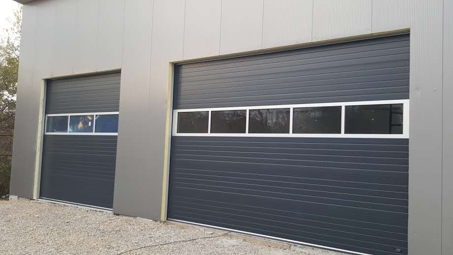Dwie szare bramy garażowe segmentowe z oknami, zamontowane w szarej hali przemysłowej. Widok z zewnątrz na żwirowe podłoże.