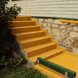 Żółte, ceramiczne schody zewnętrzne przylegają do kamiennej ściany i beżowego sidingu. Widoczna zieleń roślin i fragment metalowej barierki.
