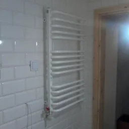 Biały grzejnik drabinkowy zamontowany na ścianie z białych, błyszczących płytek typu metro w łazience. Widoczna żółta rura instalacji gazowej pod sufitem oraz fragment drewnianej ościeżnicy drzwi.