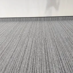 Szara wykładzina dywanowa w pionowe prążki, ułożona przy białej ścianie z czarną listwą przypodłogową.